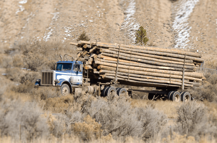 A truck with a flatbed trailer hauling tree trucks on a road through the desert.